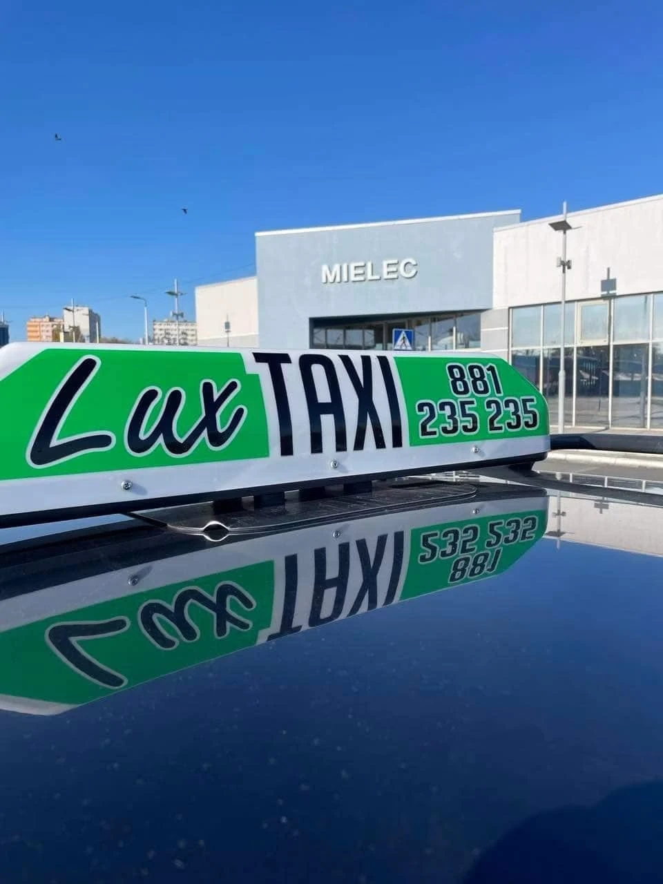 Kogut LUX Taxi na dachu samochodu przed budynkiem z napisem Mielec